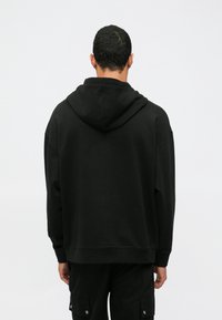 Uomo che indossa un felpa nera con cappuccio e pantaloni cargo neri, in piedi rivolto di spalle contro uno sfondo chiaro e semplice.