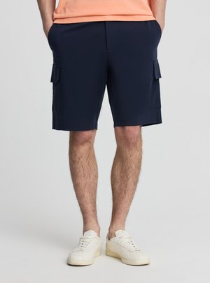 Uomo che indossa pantaloncini cargo blu navy con tasche laterali, t-shirt color pesca e sneaker bianche, in piedi davanti a uno sfondo chiaro e uniforme.
