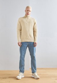Maglione beige a coste con zip, abbinato a jeans azzurri e sneakers chiare, su uno sfondo neutro con pavimento in legno.