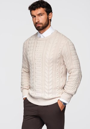 Pull en tricot beige avec un col rond et des poignets côtelés, porté par-dessus une chemise blanche à col, associé à un pantalon sombre.