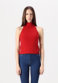 Rdeča, brezrokavna turtleneck majica iz rebraste tkanine; ima prilagojeno zasnovo in skrajšano dolžino. Nosi se z modrimi kavbojkami.