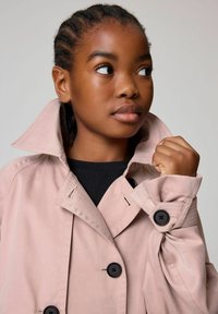 Ljusrosa trenchcoat med hög krage, svarta knappar och strukturerade ärmar. Bärs över en svart topp som visar en ren design.