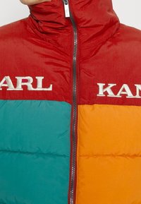 Färgblockerad puffjacka med röda, turkosa och orange sektioner. Materialet har en slät, glänsande yta med hög krage och dragkedja.