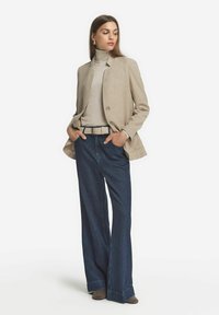 Beige kavaj, ljus polokrage, mörka vida jeans och bruna klackar. Kavajen har en strukturerad yta och enkel knappstängning.