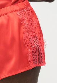 Κόκκινα σατέν σορτς με ελαστική μέση και κάθετη λουλουδάτη δαντέλα στο πλαϊνό μέρος, σε απλό φόντο.