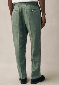 Grüne Leinenhose mit geradem Schnitt, versehen mit zwei Gesäßtaschen mit Knopfverschluss und einem flachen Bund. Getragen mit dunklen Schuhen.