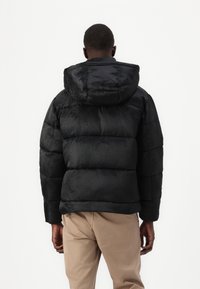 Melnā pūkainā jaka ar kapuci, ar izšūtu dizainu, maigu logo rakstu un elastīgām piedurknēm. Tekstūra izskatās gluda un spīdīga.