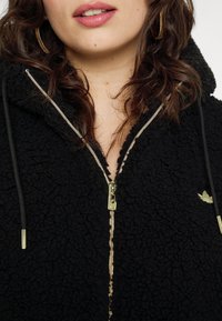 Svart fleece zip-up jacka med en texturerad yta, guld dragkedja och snörningsdetaljer. Litet logo på bröstet.