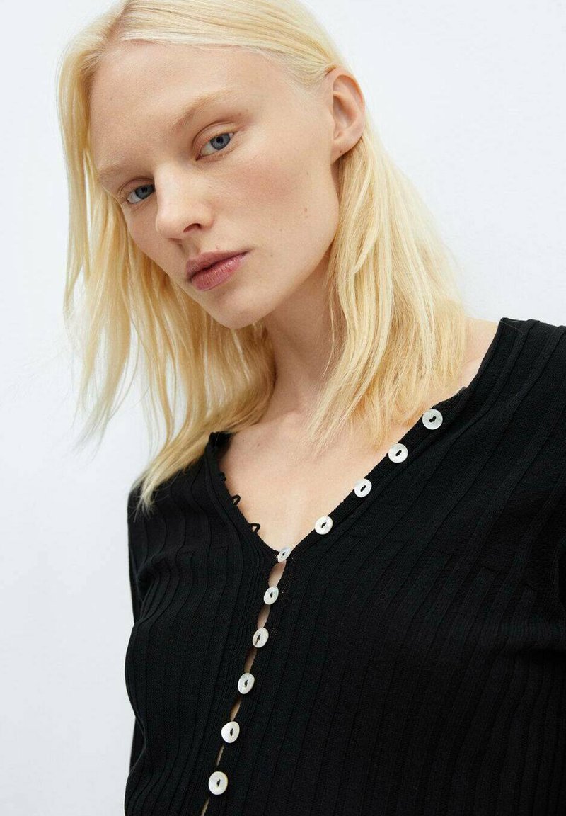 Cardigan nero a coste con scollo a V, caratterizzato da bottoni bianchi a cerchio lungo la parte frontale. Il materiale sembra morbido e strutturato.