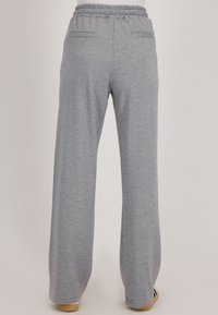 Achteraanzicht van een persoon die losse lichtgrijze trainingsbroek met elastische tailleband en twee achterzakken draagt, staand op een lichte achtergrond.