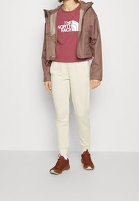 Bruine cropped jas met capuchon, bordeaux t-shirt met witte logo, crèmekleurige trainingsbroek en bruine sportschoenen met groene zolen.