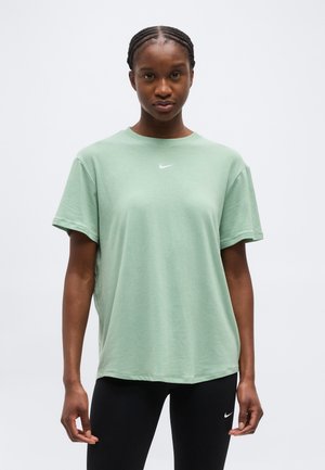 Kvinne med flettehår iført en lys grønn Nike T-skjorte og svarte treningsbukser, står mot en ensfarget hvit bakgrunn.