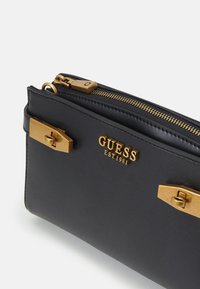 Schwarze Clutch aus synthetischem Material mit goldfarbenen Beschlägen, ausgestattet mit einem Reißverschluss und dem Logo "GUESS EST.1981." Die Beschläge umfassen zwei dekorative Verschlüsse.