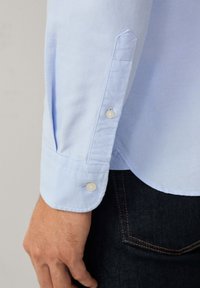 Polsino di camicia azzurro chiaro a manica lunga con bottoni bianchi sul braccio di una persona, accanto a jeans di denim scuro con cuciture arancioni.