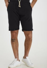Shorts en coton noir avec taille élastique à cordon de serrage, ourlets roulés et poches latérales. Associés à des baskets blanches sur un fond clair.