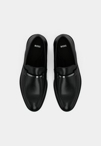 Et par svarte skinn herre loafers med metallspenne-detall, sett ovenfra mot en hvit bakgrunn, innersåler merket "BOSS."