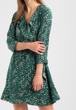 Frau in einem grünen Blumenkleid mit Rüschen am Ausschnitt und Saum, die mit verschränkten Armen posiert.