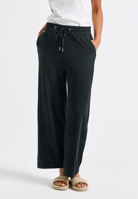 Svarta vida byxor i mjuk tyg, med elastisk midja och dragsko samt sidofickor. Bärs med ljusa sandaler.