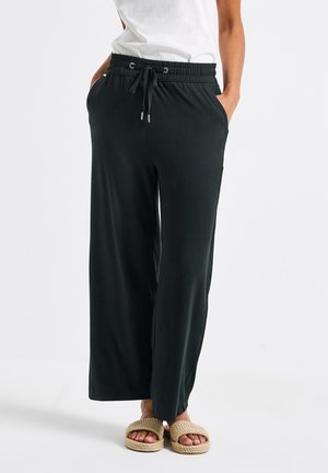 Pantaloni larghi neri realizzati in morbido tessuto, con vita elastica e cordino e tasche laterali. Indossati con sandali di colore chiaro.