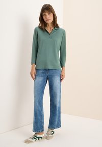 Polo verde a maniche lunghe con colletto, tre bottoni, abbinato a jeans a gamba larga azzurri e sneakers grigie con dettagli verdi.