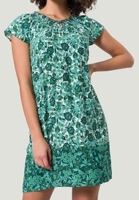 Robe courte florale verte avec des manches courtes, décolleté froncé et un design fluide. Présente des nuances variées de vert et des motifs complexes.
