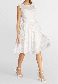 Robe blanche avec un corsage transparent et une jupe en couches ressemblant à des plumes. Associée à des talons aiguilles blancs ornés de rubans.
