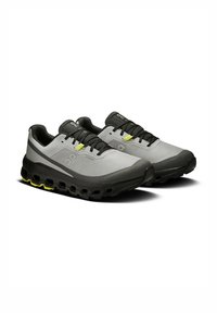 Graue Laufschuhe mit schwarzen Akzenten, strukturiertem Stoff und einer klobigen Sohle, die sichtbare Dämpfungslöcher und ein leuchtend gelbes Detail aufweist.
