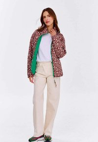 Brązowa kurtka puchowa w kropki z zielonym wnętrzem, z zapięciem na zamek i koralikowym sznurkiem. Połączona z kremowymi spodniami i wzorzystymi butami.