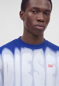 Jeune homme aux cheveux courts et bouclés portant un sweat-shirt tie-dye bleu et blanc avec un logo Diesel rouge sur la poitrine sur fond gris.