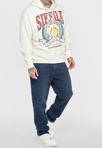White hoodie with red and green graphic text, paired with dark blue jeans and white sneakers. Hood tightly fitted with drawstrings.