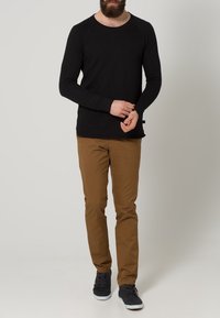 Man som bär en svart långärmad skjorta, bruna byxor och mörka sneakers som står mot en enkel grå bakgrund.