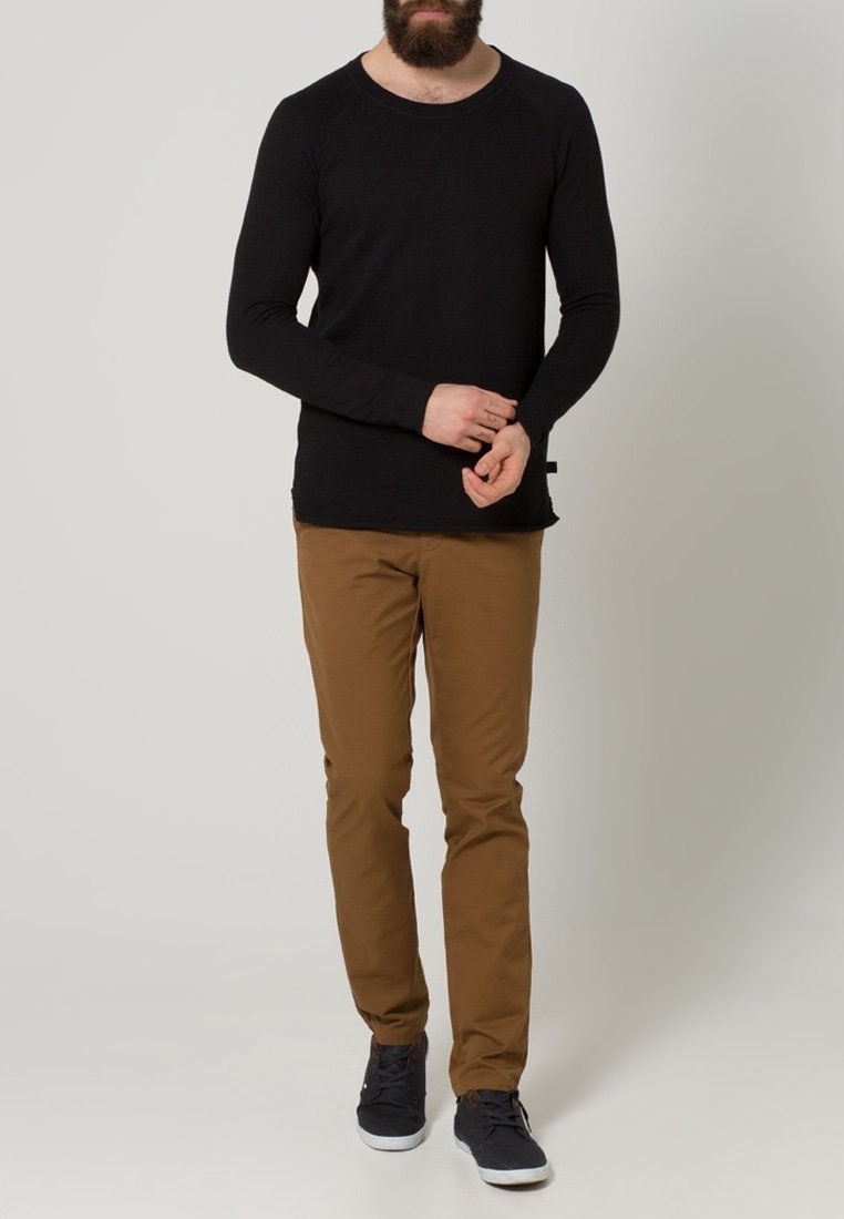 Man som bär en svart långärmad skjorta, bruna byxor och mörka sneakers som står mot en enkel grå bakgrund.