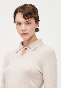 Beige neule, jossa on kauluksellinen kaula, etuosassa pieni avaus. Neule on valmistettu pehmeästä materiaalista, jossa on hienovarainen tekstuuri.