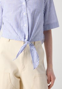 Blå og hvitstripet skjorte med knapp og knyttet front, kombinert med beige høytlivshorts med frontlommer.