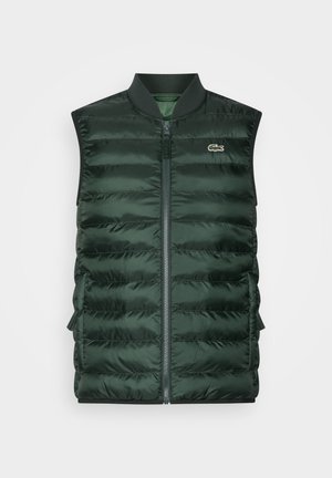 Groene puffervest over een lichtblauwe geblokte shirt, gecombineerd met donkergroene broek en witte sneakers. De pet is marineblauw en het materiaal lijkt glad.