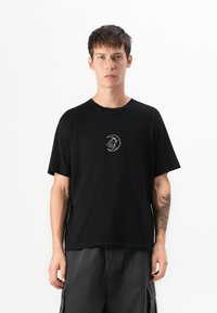 Junger Mann mit kurzen, dunklen Haaren, der ein schwarzes T-Shirt mit einem weißen Mond- und Wolkendesign trägt, vor einem schlichten weißen Hintergrund stehend.
