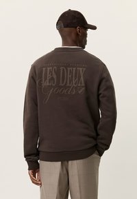 Καφέ φούτερ με στρογγυλή λαιμόκοψη και μανσέτες με ριμπ ύφανση. Διαθέτει μεγάλο, τυπωμένο λογότυπο σε τονικό χρώμα στο πίσω μέρος. Υλικό από βαμβακερό μείγμα.