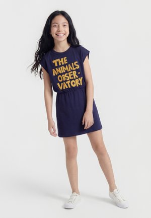 Ragazza sorridente, indossa un vestito blu navy con testo giallo "THE ANIMALS OBSERVATORY" e sneakers bianche, in piedi contro uno sfondo bianco uniforme.