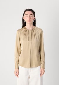 Blusa a maniche lunghe in morbido satin color beige chiaro, con dettaglio di scollatura arricciata e linea dritta, abbinata a pantaloni bianchi.