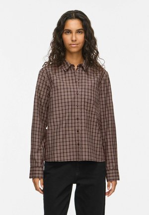 Femme aux cheveux bouclés et foncés portant une chemise à carreaux marron boutonnée et un pantalon noir, debout devant un fond clair uni.