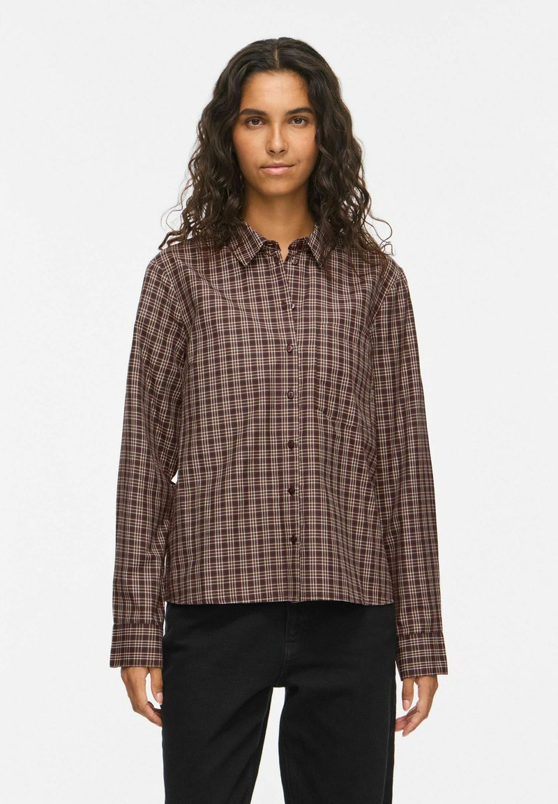 Femme aux cheveux bouclés et foncés portant une chemise à carreaux marron boutonnée et un pantalon noir, debout devant un fond clair uni.