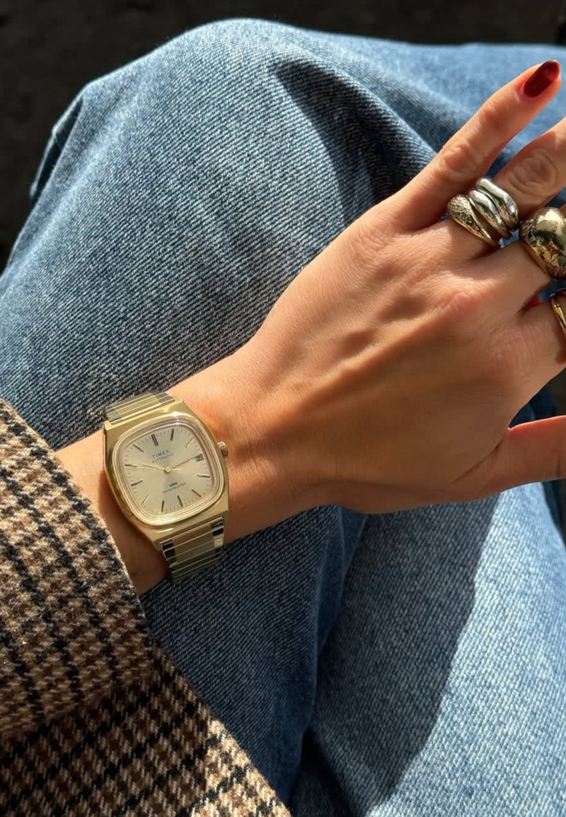 Silver wristwatch with a rectangular face and an embossed texture, set on a metal bracelet. Hand adorned with multiple silver rings.