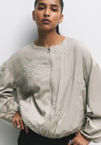 Giacca bomber grigia chiaro con ricami floreali, chiusura con zip e polsini elasticizzati. Tessuto liscio simile alla seta con vestibilità rilassata.