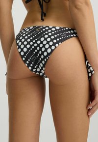 Čierne bikiny so vzorom čiernych bodiek v bielej farbe, so šnúrkami na bokoch a malým kovovým logom. Hladká textúra.