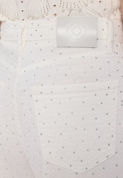 Weiße Denim-Jeans, verziert mit kleinen silbernen Nieten, mit strukturiertem Oberflächenmaterial und einem glatten weißen Lederschild am Bund mit dekorativem Design.