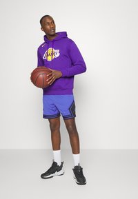 Felpa viola con il logo "Los Angeles Lakers", pantaloni corti in rete blu con dettagli neri e scarpe da ginnastica nere. Palla da basket tenuta nella mano destra.