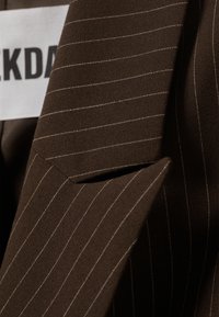 Brązowa tkanina w paski z teksturowanym wykończeniem, z klapami w kształcie litery V i białą metką z napisem "ALDKDA."