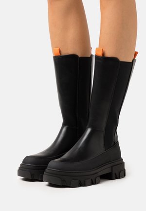 Hoge zwarte leren laarzen met grove, geribbelde zolen en oranje trekbanden, gedragen op blote benen tegen een witte achtergrond.