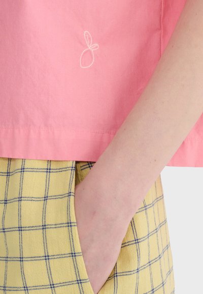 Un bras à la peau claire repose dans la poche d'un pantalon à carreaux jaunes, associé à un tissu rose orné d'un petit motif blanc brodé.