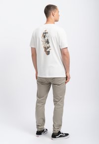 Witte katoenen t-shirt met een skateboardafbeelding op de achterkant, gecombineerd met beige broek en zwarte sneakers. Eenvoudig ontwerp en een relaxte pasvorm.